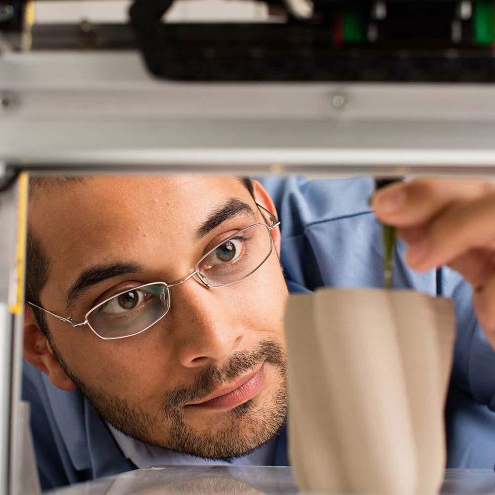 一名戴眼镜的男学生正在调整3D打印机，打印出部分完成的曲线米色花瓶