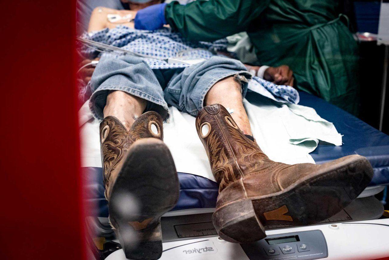 躺在医院病床上的老人, 由一个穿着绿色长袍，戴着蓝色手套的医务人员照料. 病人的牛仔裤被拉到膝盖以上, 破旧的牛仔靴挂在床沿上.