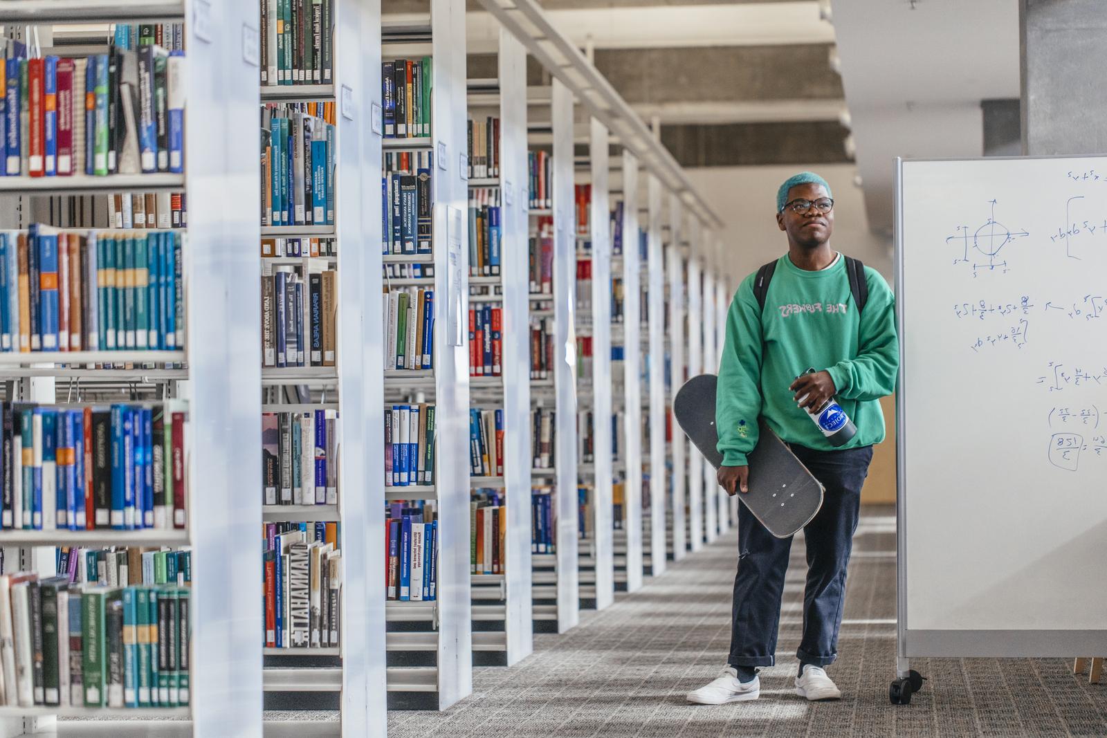 一个穿着绿色运动衫、拿着滑板的年轻人靠在图书馆书架旁边的一块白板上
