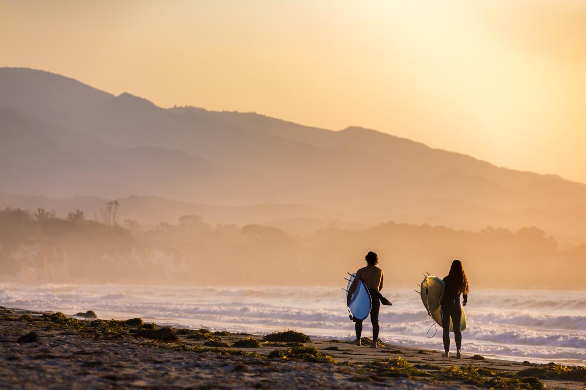 两个冲浪者在日落时沿着海滩散步，背景是山脉