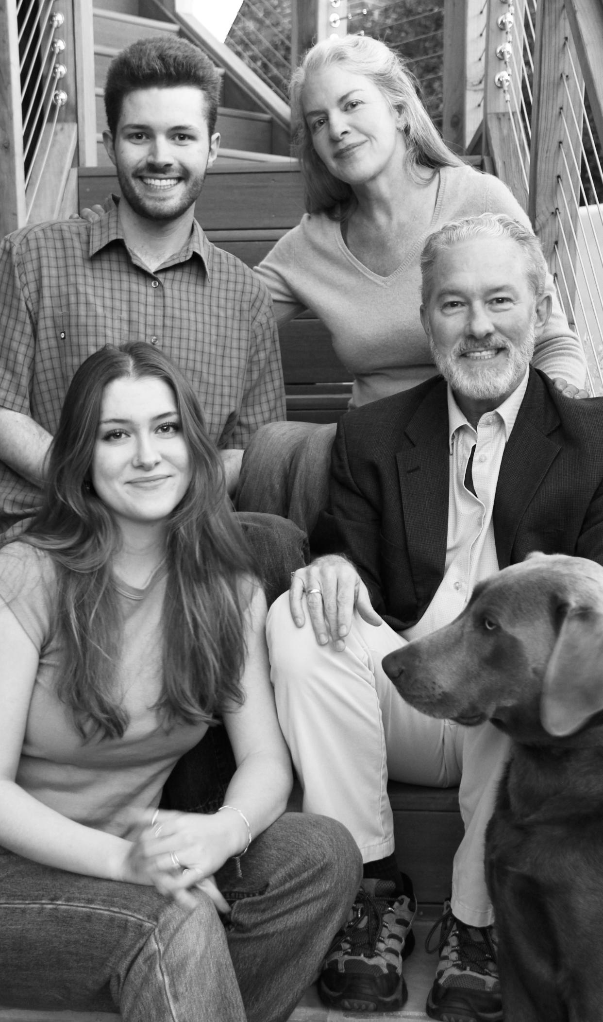 A black and white photo of Rich Lyons and his family seated on stairs with his daughter seated next to him; wife seated above; son seated next to her; and Weimaraner dog poking his head in 