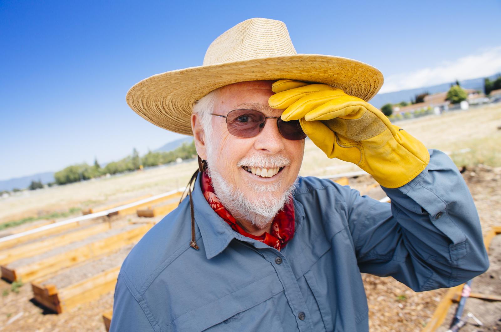 戴着墨镜的白胡子男人, 蓝色衬衫, 黄色的手套, 草帽和红手帕擦着额头，在农田里微笑.