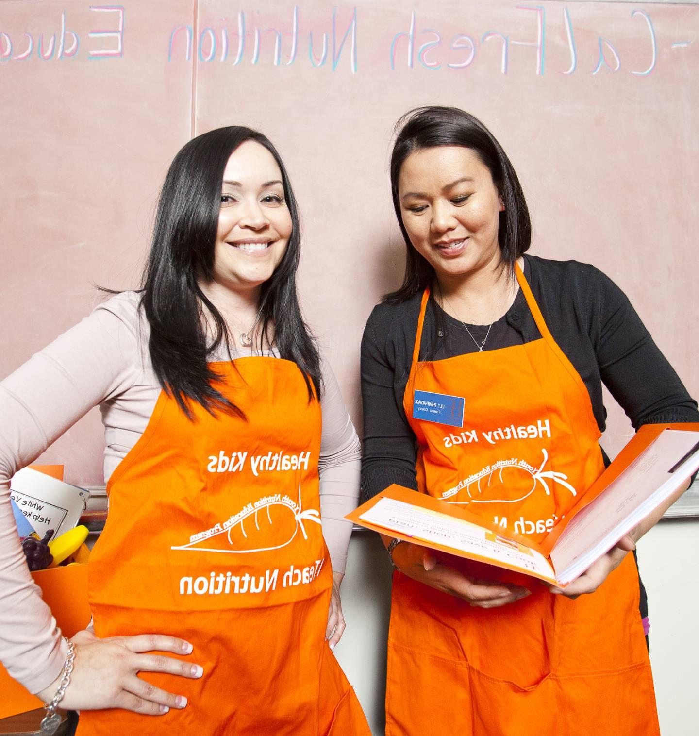 Two women in orange aprons one holding a book