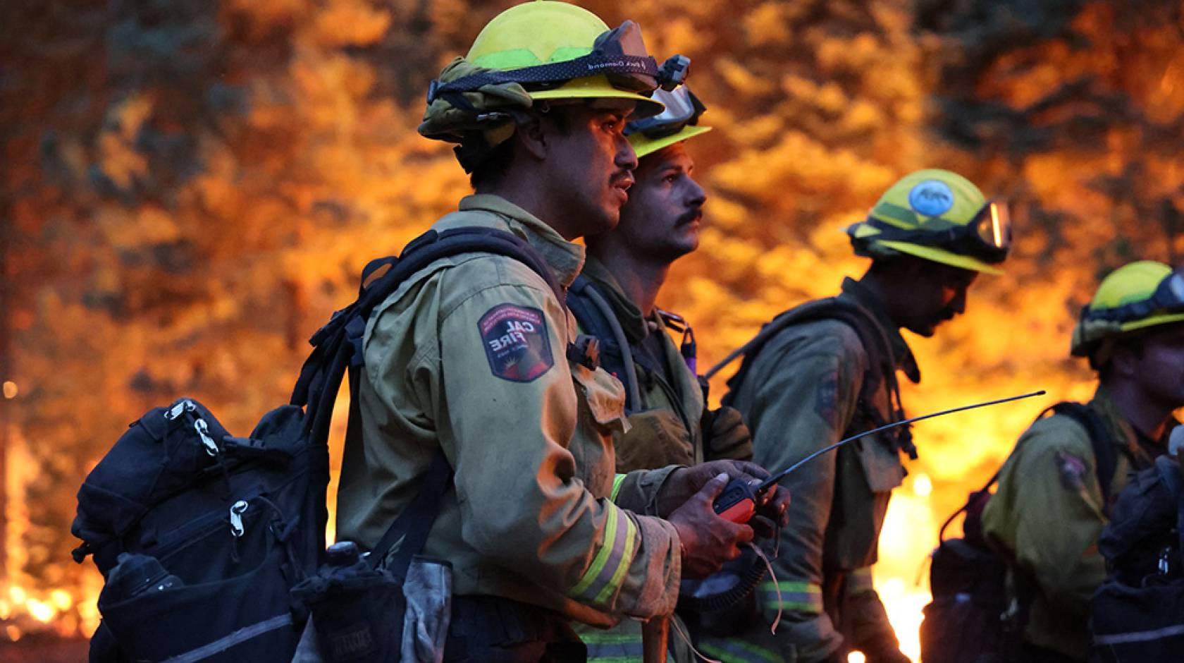 四名消防员站在火焰的背景下. 现场很黑，两名年轻的男性消防员拿着一个带长天线的装置