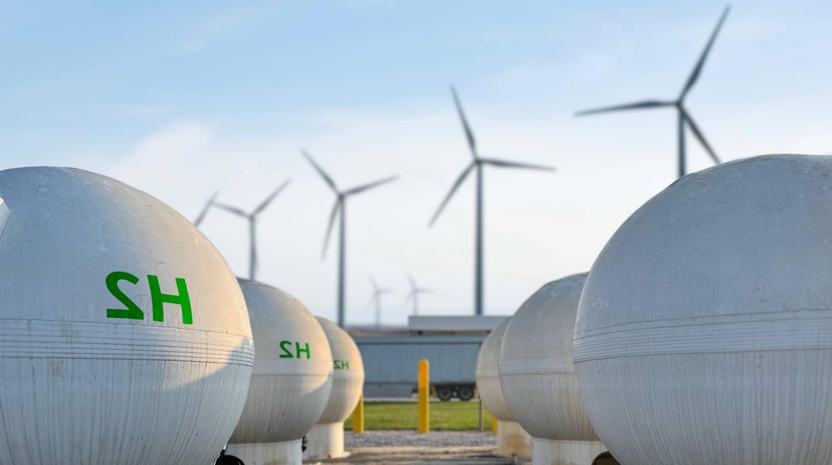两排白色的球形罐，上面用绿色印着H2, 背景是四个风力涡轮机, 湛蓝的天空