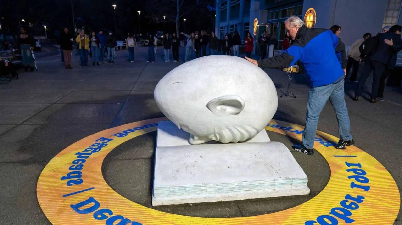 In the front of the photo, a man touches an egghead (a humorous but blank-looking human face shaped like an egg) which is faced down on a concrete open book; in the back of the photo, a line of people outside a museum at night
