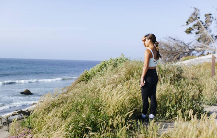 一个穿着运动服的年轻女子站在长满草的悬崖上，俯瞰着海滩, shading her eyes from the sun