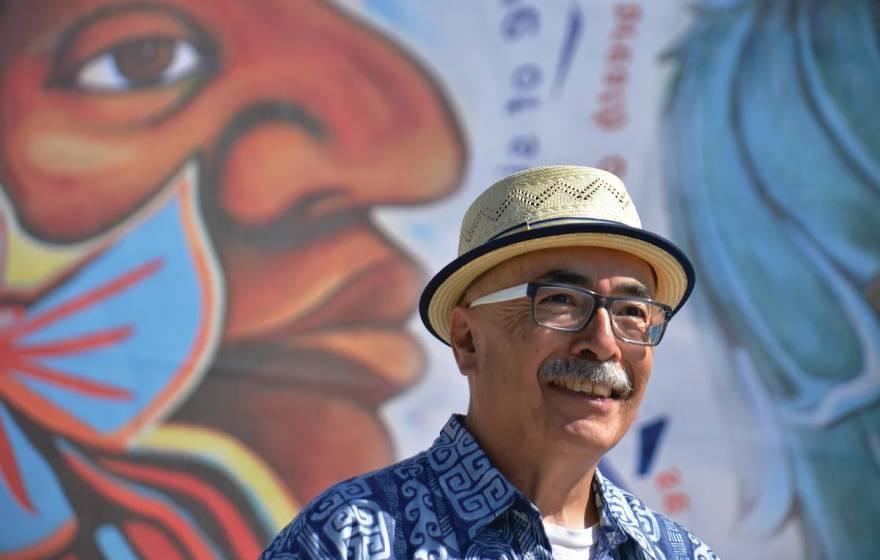 一位留着小胡子、戴着眼镜、戴着时髦帽子的拉丁裔老人站在一幅彩色壁画前
