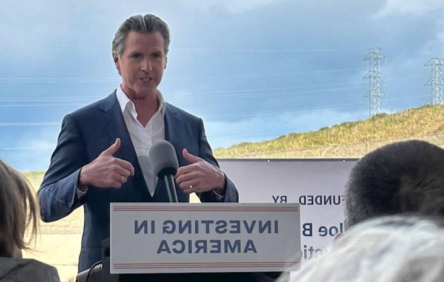 Governor Newsom speaking at a podium that says Investing in America, with countryside behind him