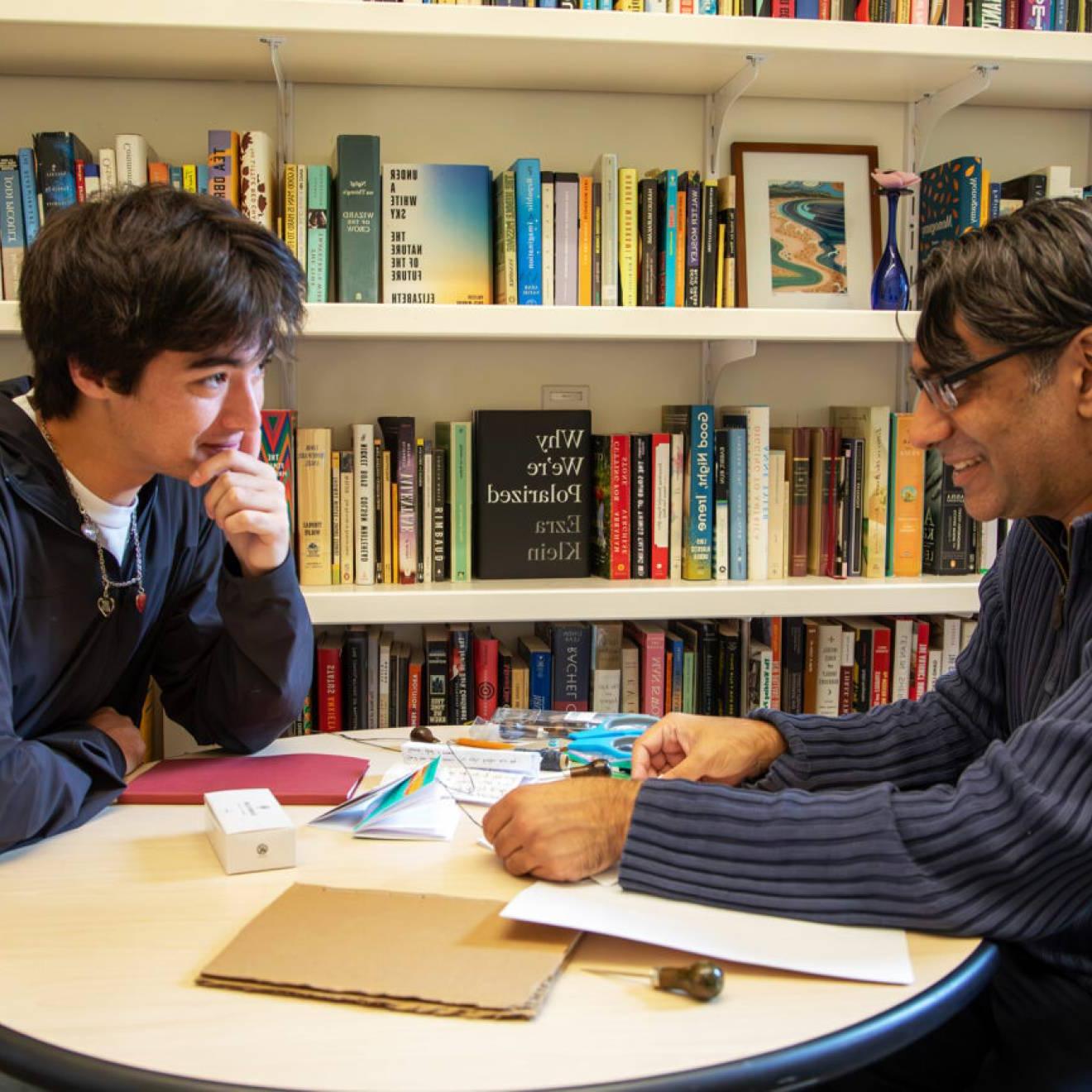 一个教授年龄的男人和一个学生年龄的男人坐在一个书架前的圆桌旁, 面对面讨论. 