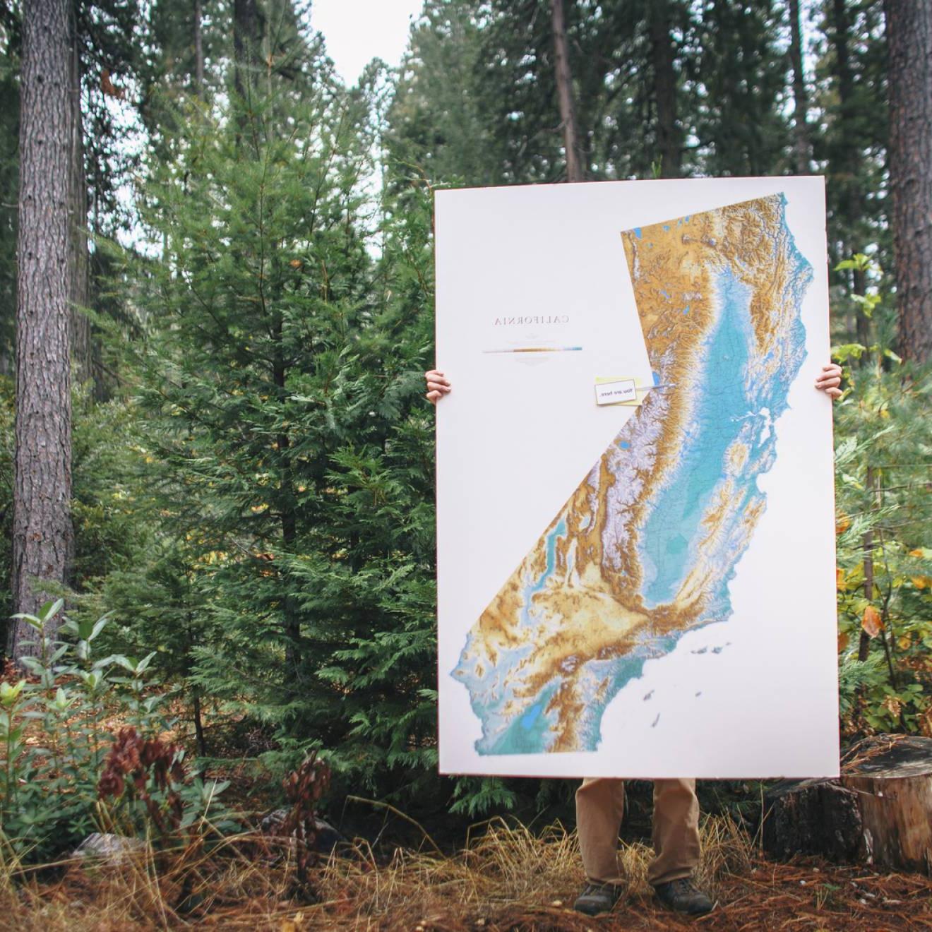 A person stands in a forest, holding a big map of California, which covers all but their legs and hand. 