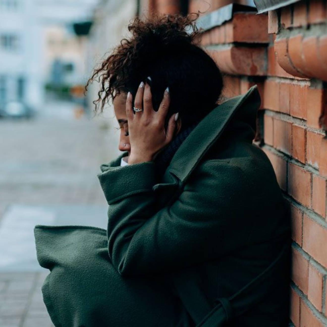 A young Black woman crouches against a brick wall holding her head
