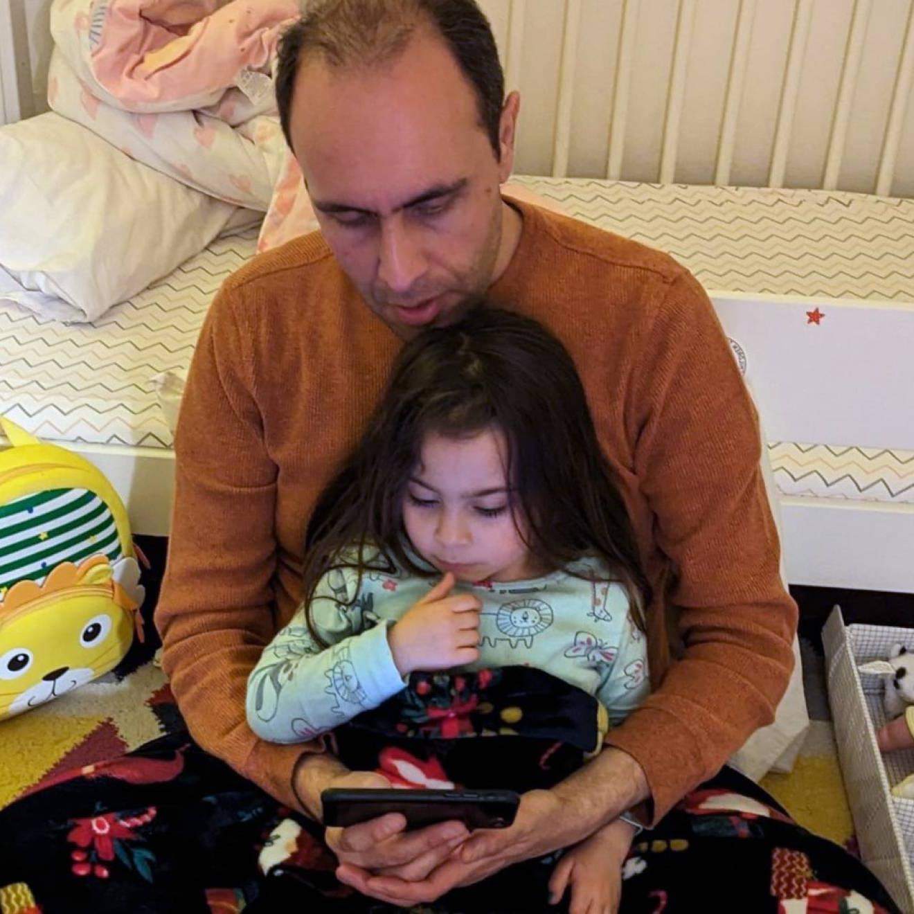 一个秃顶的男人抱着一个年幼的女儿，他们一起阅读，看着屏幕