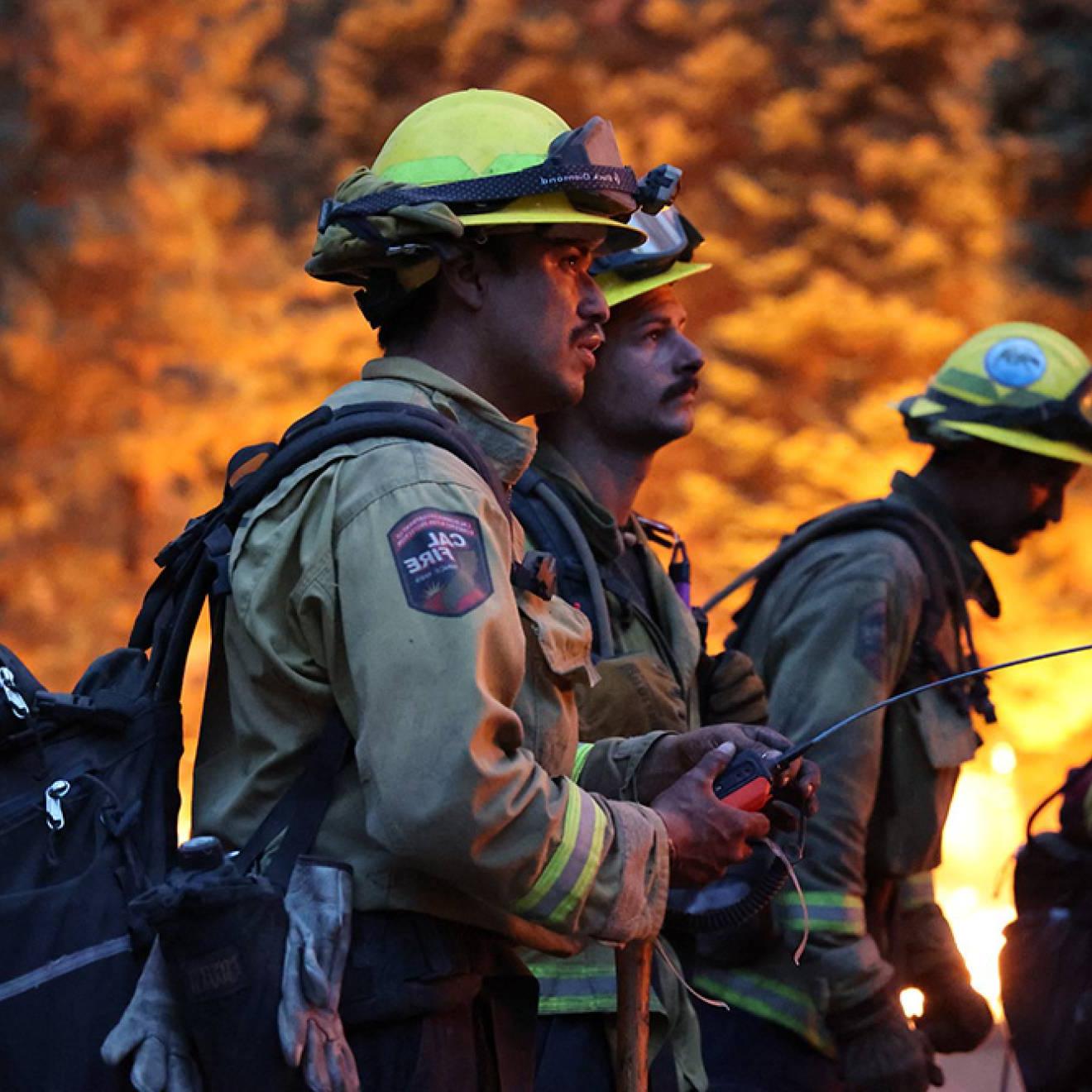 Four firefighters stand against a backdrop of flames. 现场很黑，两名年轻的男性消防员拿着一个带长天线的装置
