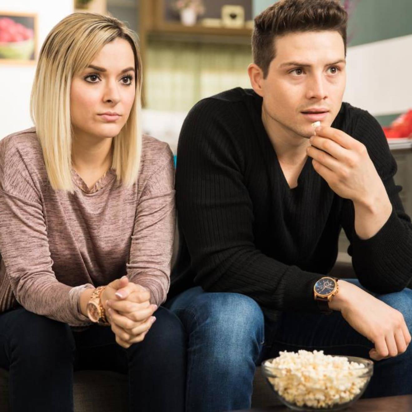 A white man and a white woman watching TV together, looking somber