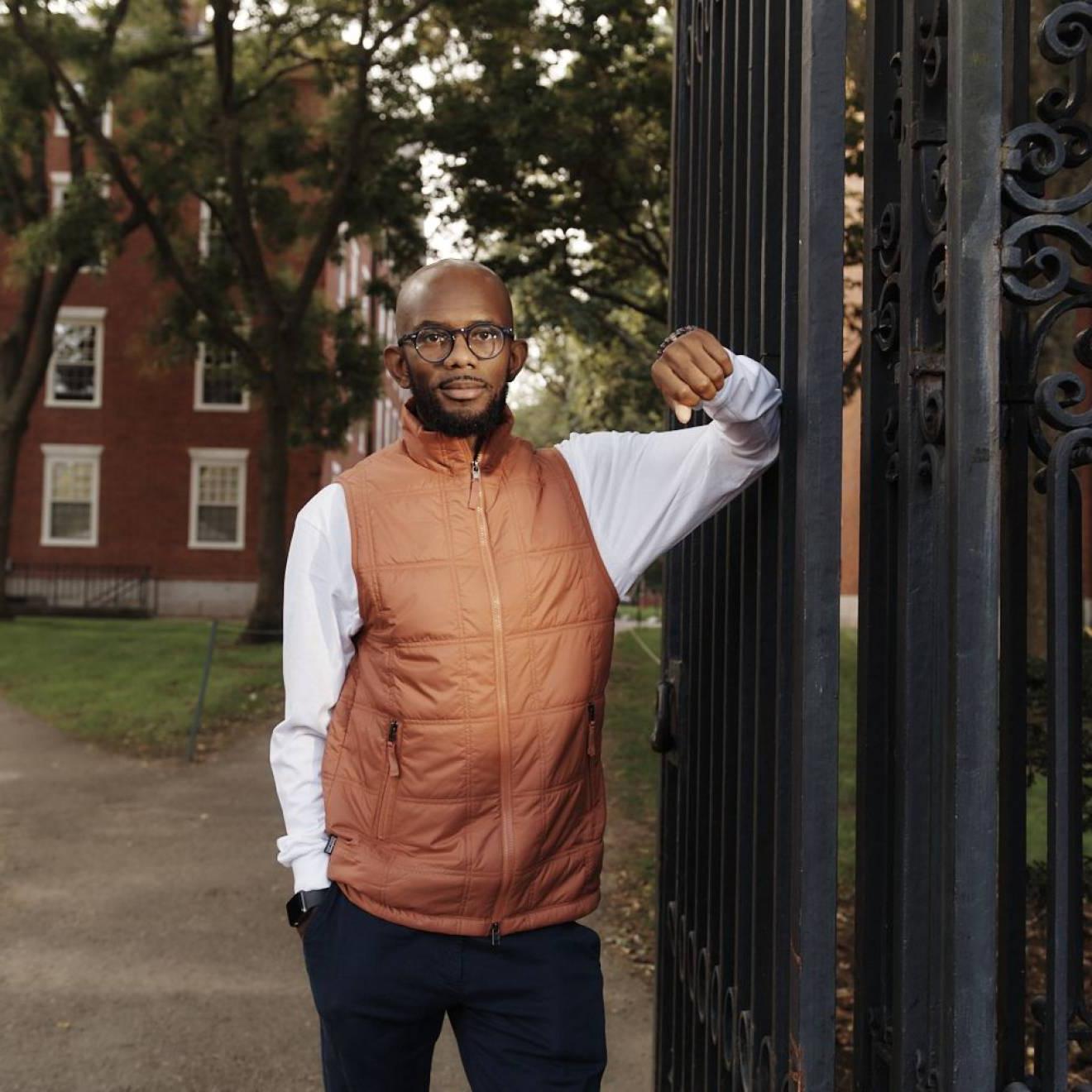 身穿橙色背心、戴着眼镜的年轻黑人站在哈佛校园的一扇大门前
