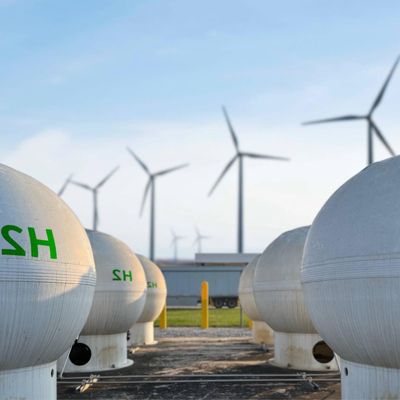 两排白色的球形罐，上面用绿色印着H2, 背景是四个风力涡轮机, 湛蓝的天空