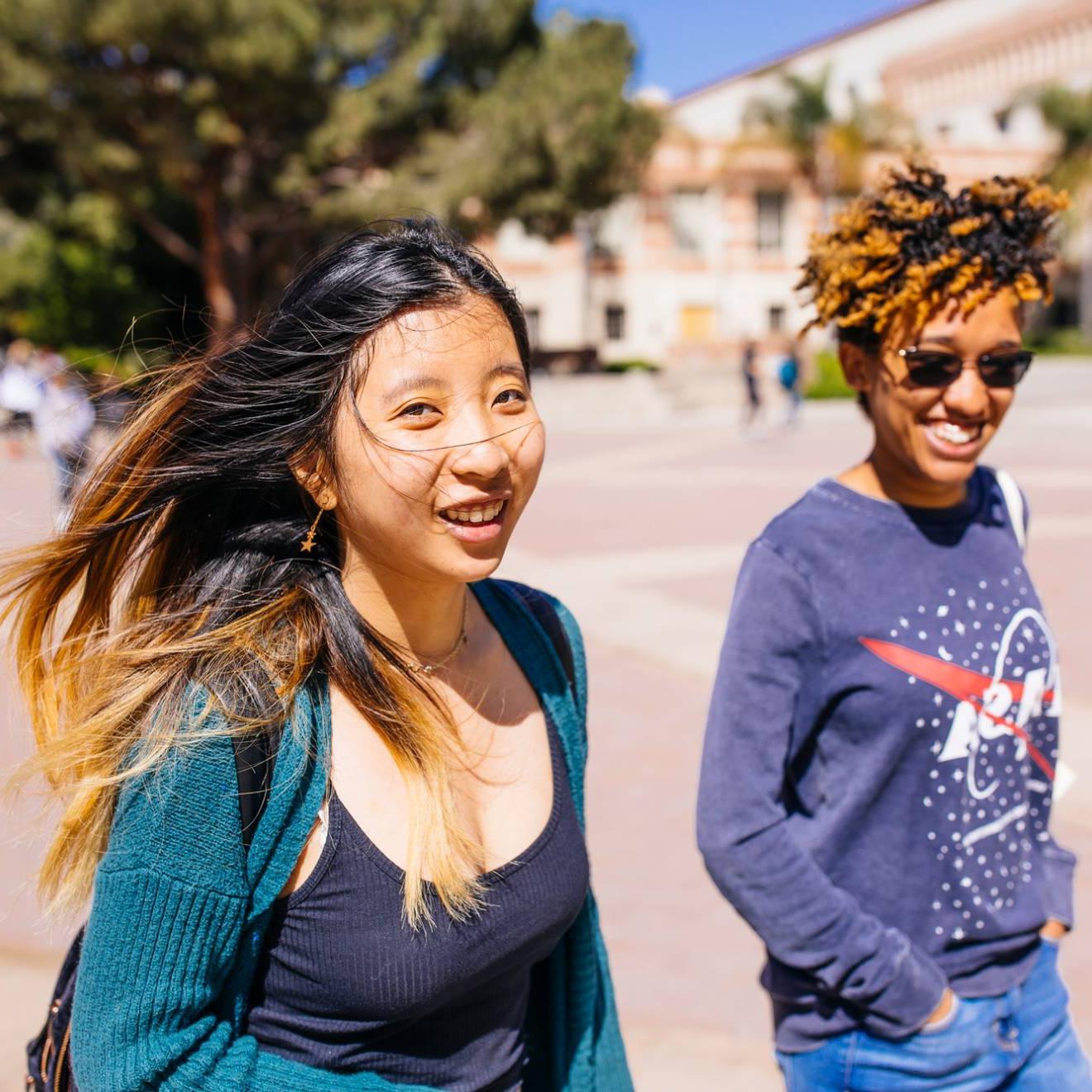 一个穿着NASA运动衫的年轻黑人女性和一个穿着绿色开襟羊毛衫的年轻亚洲女性微笑着走在校园里
